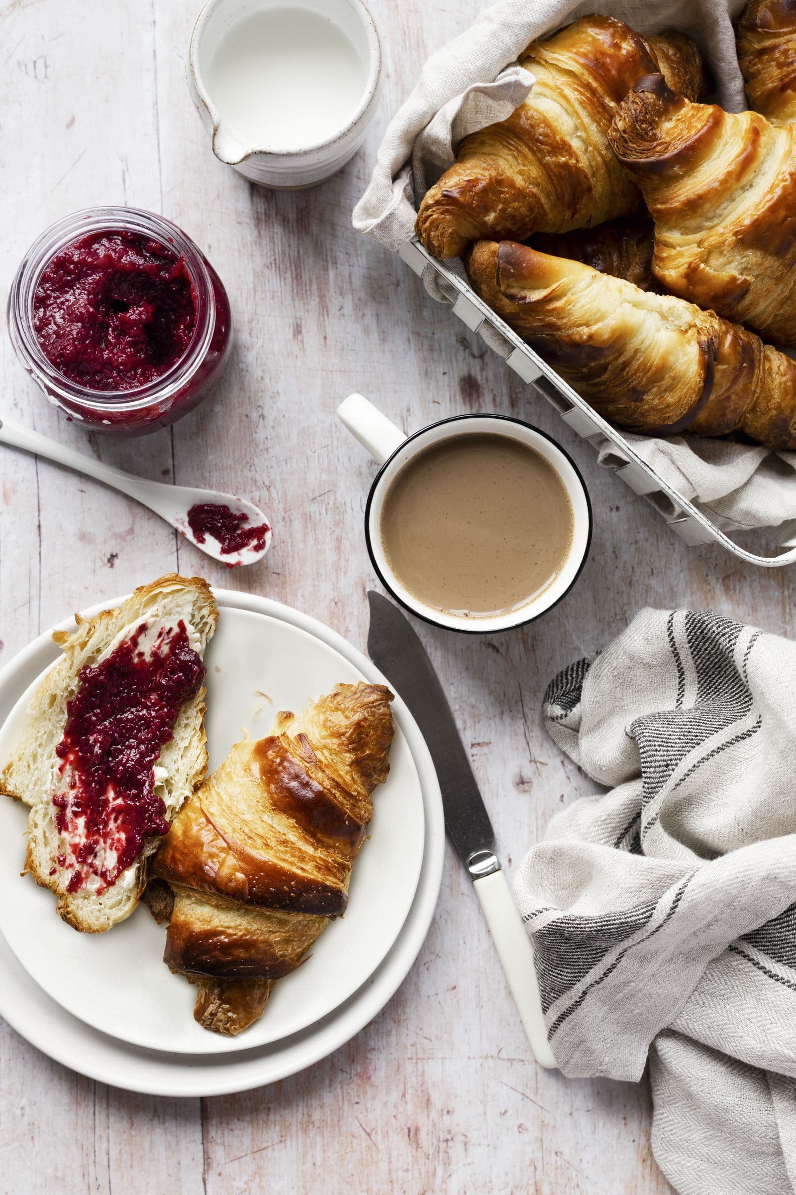 Croissants im Korb, eine Tasse Milchkaffee und ein Brötchen mit roter Marmelade