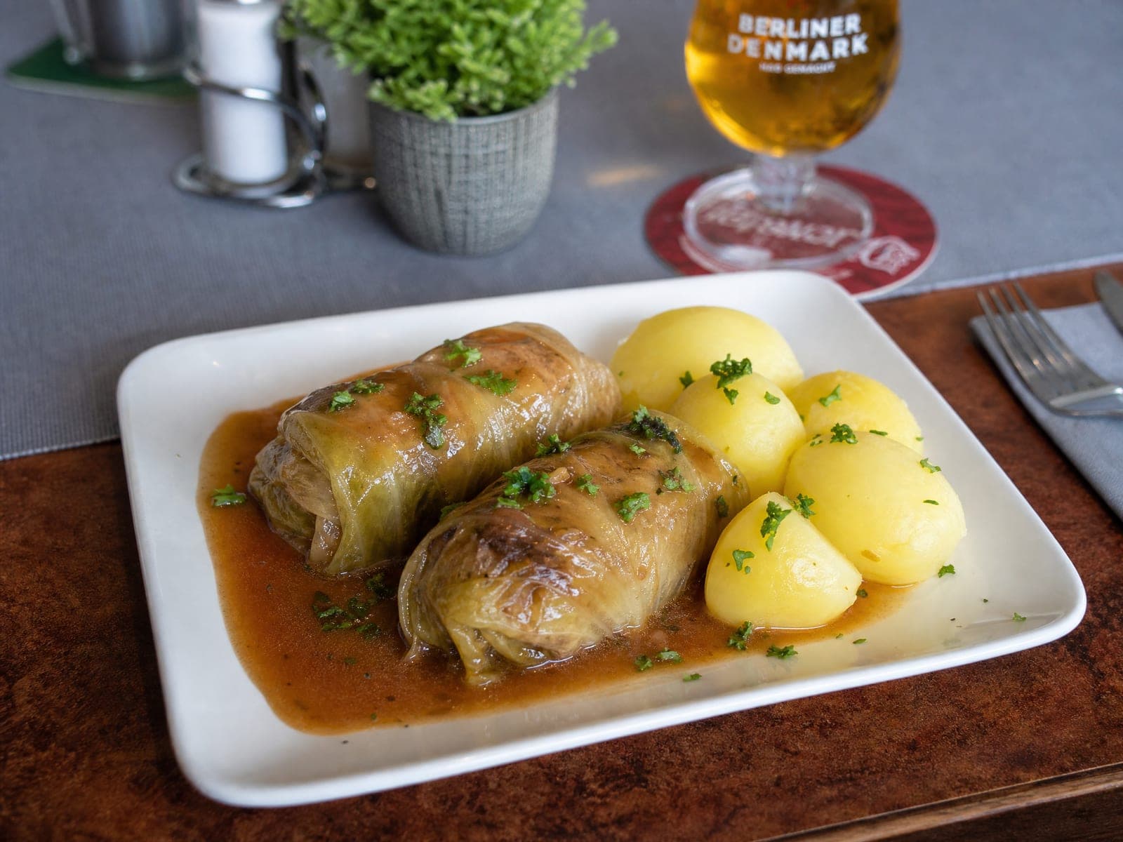Zwei Kohlrouladen in brauner Soße mit Salzkartoffeln und Petersilie, dahinter ein Glas Bier